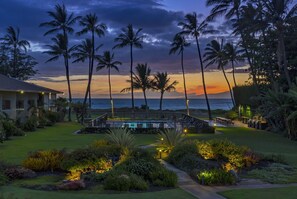 A pool area with a view of the ocean, surrounded by palm trees and well-lit pathways. - Waiohuli Beach Hale C212 (Kihei)