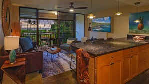 A modern kitchen with a large island, dining area with chairs, and a view of the ocean through large windows. - Waiohuli Beach Hale C212 (Kihei)