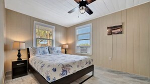 A bedroom with a bed, two nightstands, a ceiling fan, and two windows with blinds. - Stones Throw Beachside Bungalow 1 ON THE Beach (Indian Rocks Beach)