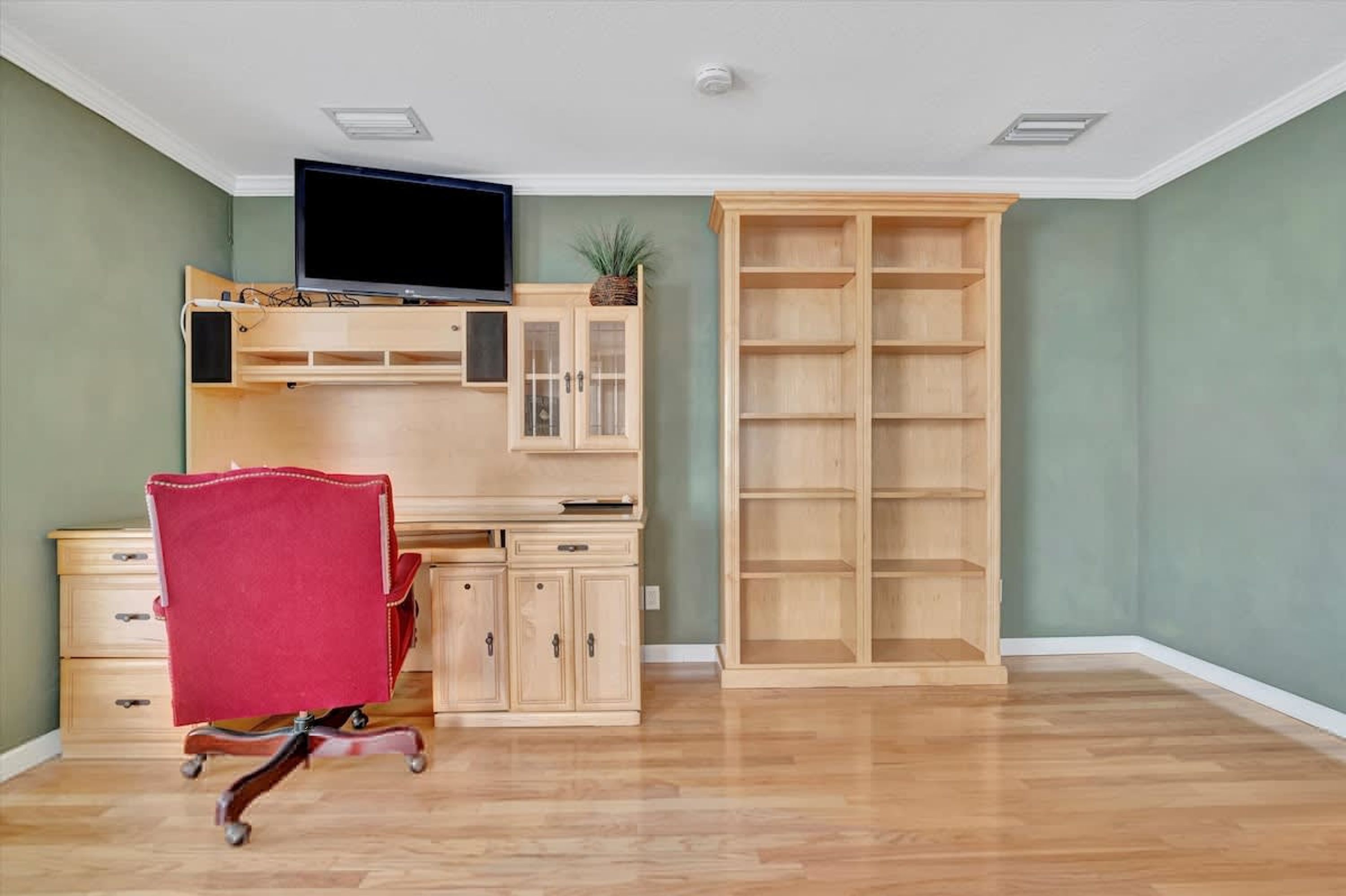 Une pièce avec un bureau en bois, une chaise rouge, un téléviseur et une grande étagère en bois.