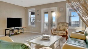 Een woonkamer met een televisie, een plafondventilator, een bank, een fauteuil en een openslaande deur naar een balkon.