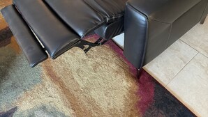 Un sillón de cuero negro reclinable con reposapiés, dos cojines color beige y una mesita auxiliar con una lámpara.
