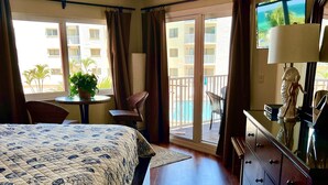 Habitación de hotel con cama, tocador, silla, mesa con planta y vista a una piscina y edificios.