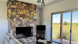 A living room with a stone fireplace, a sofa, a television on a stand, and a large sliding glass door leading to an outdoor area. - La Colina by Carmelodays (Carmelo)