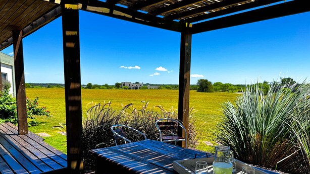 A wooden pergola with a table and chairs, a tray with glasses and a pitcher, and a view of a green field and a building in the distance. - La Colina by Carmelodays (Carmelo)