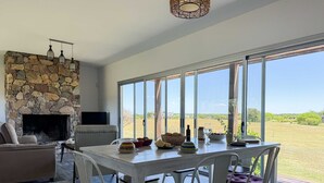 A modern living room with a stone fireplace, a flat-screen TV, and a dining area with a view of the countryside. - La Colina by Carmelodays (Carmelo)