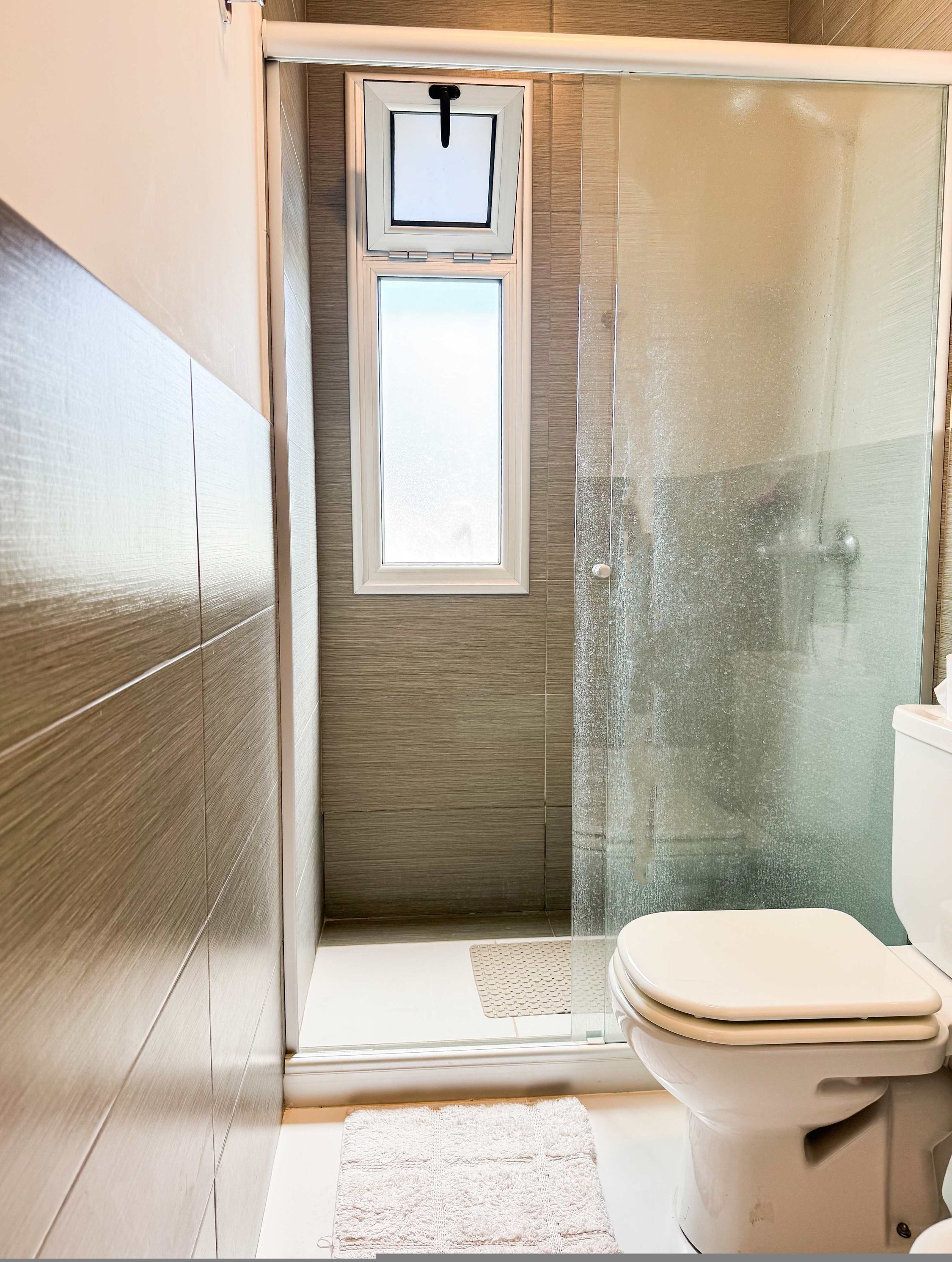 A modern bathroom with a glass shower enclosure, a toilet, and a small window.