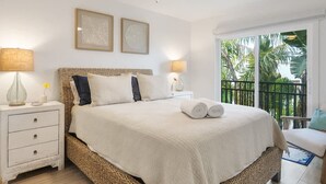 Un dormitorio con cama, mesitas de noche, lámparas y un ventilador de techo. Tiene un balcón con vista a palmeras.