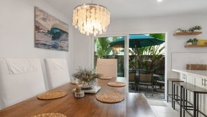 Un comedor con mesa de madera, individuales tejidos, una lámpara colgante y vista a un patio exterior con sillas y sombrillas.