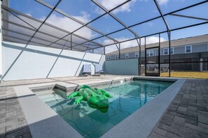 Ein Swimmingpool mit einem grünen, aufblasbaren Krokodil, einer überdachten Terrasse und Blick auf ein Wohngebäude im Hintergrund.