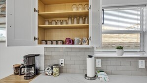 Une cuisine avec des placards ouverts où sont exposés des verres, une machine à café et une fenêtre avec des stores.