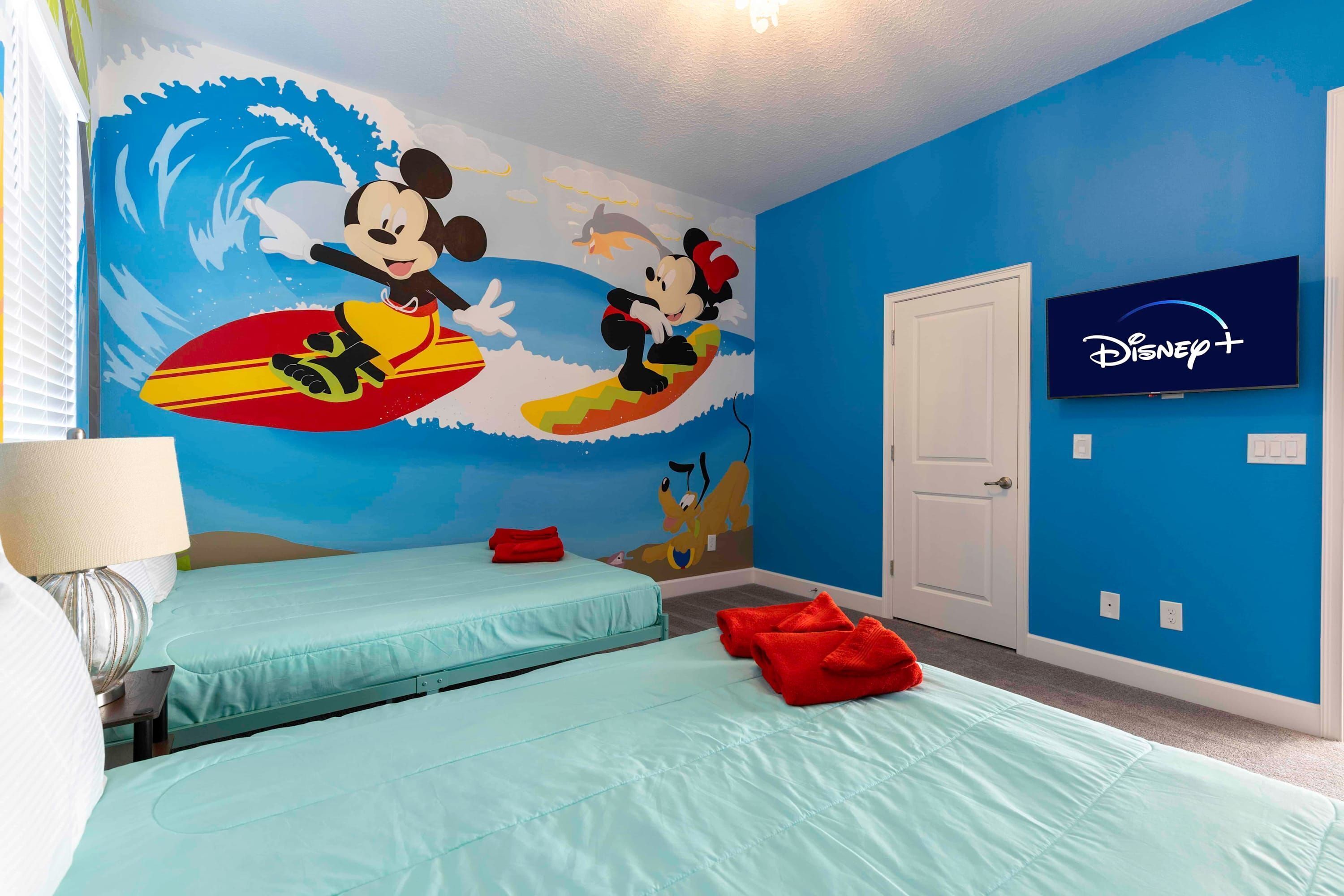 Una habitación con dos camas, un mural en la pared de Mickey y Minnie Mouse surfeando, un televisor de Disney+ y una puerta.