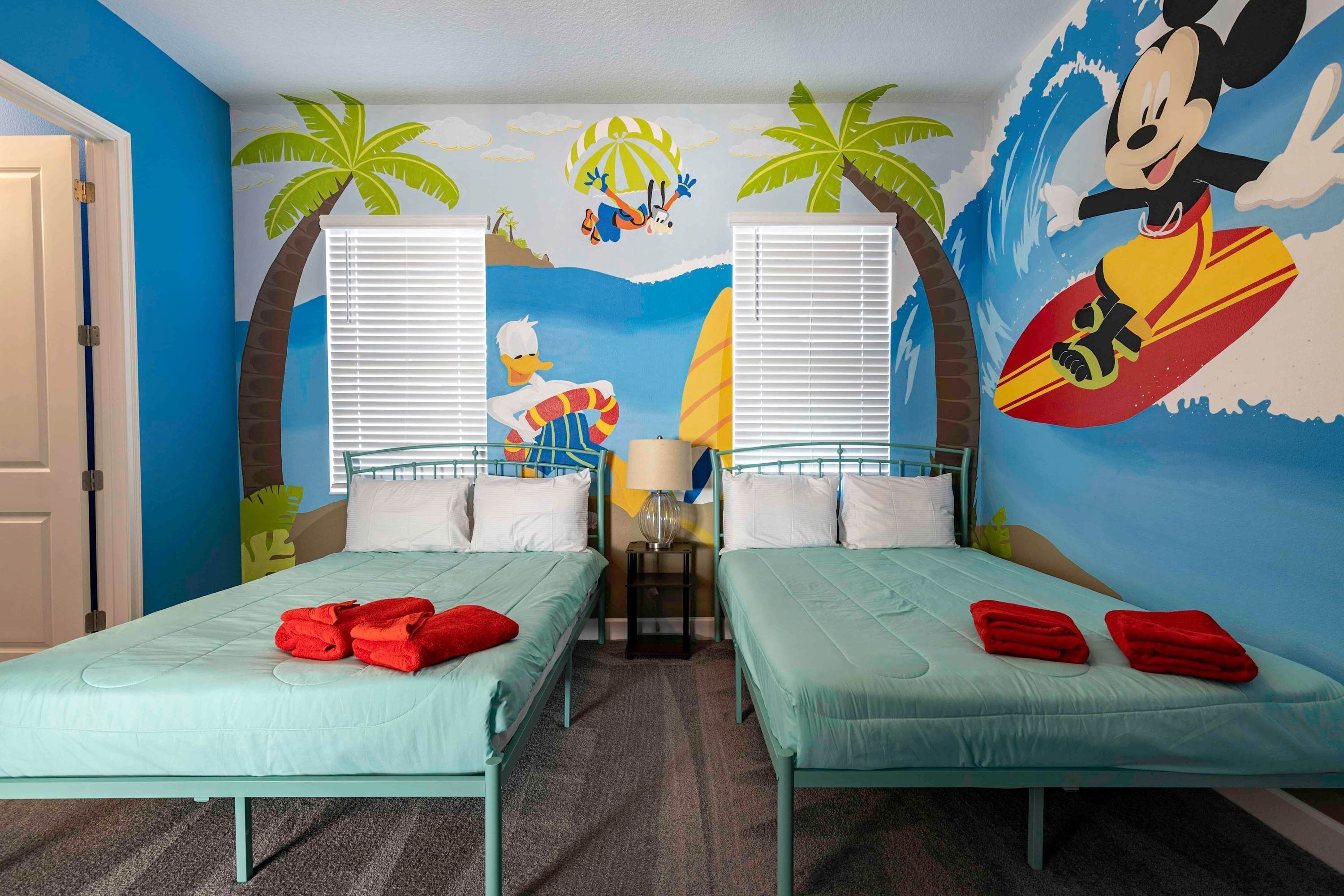 Ein Zimmer mit zwei Betten, jedes mit einer hellblauen Matratze und roten Handtüchern. An den Wänden befinden sich Wandgemälde mit Palmen und ein Wandgemälde mit einem Surfbrett von Mickey Mouse.