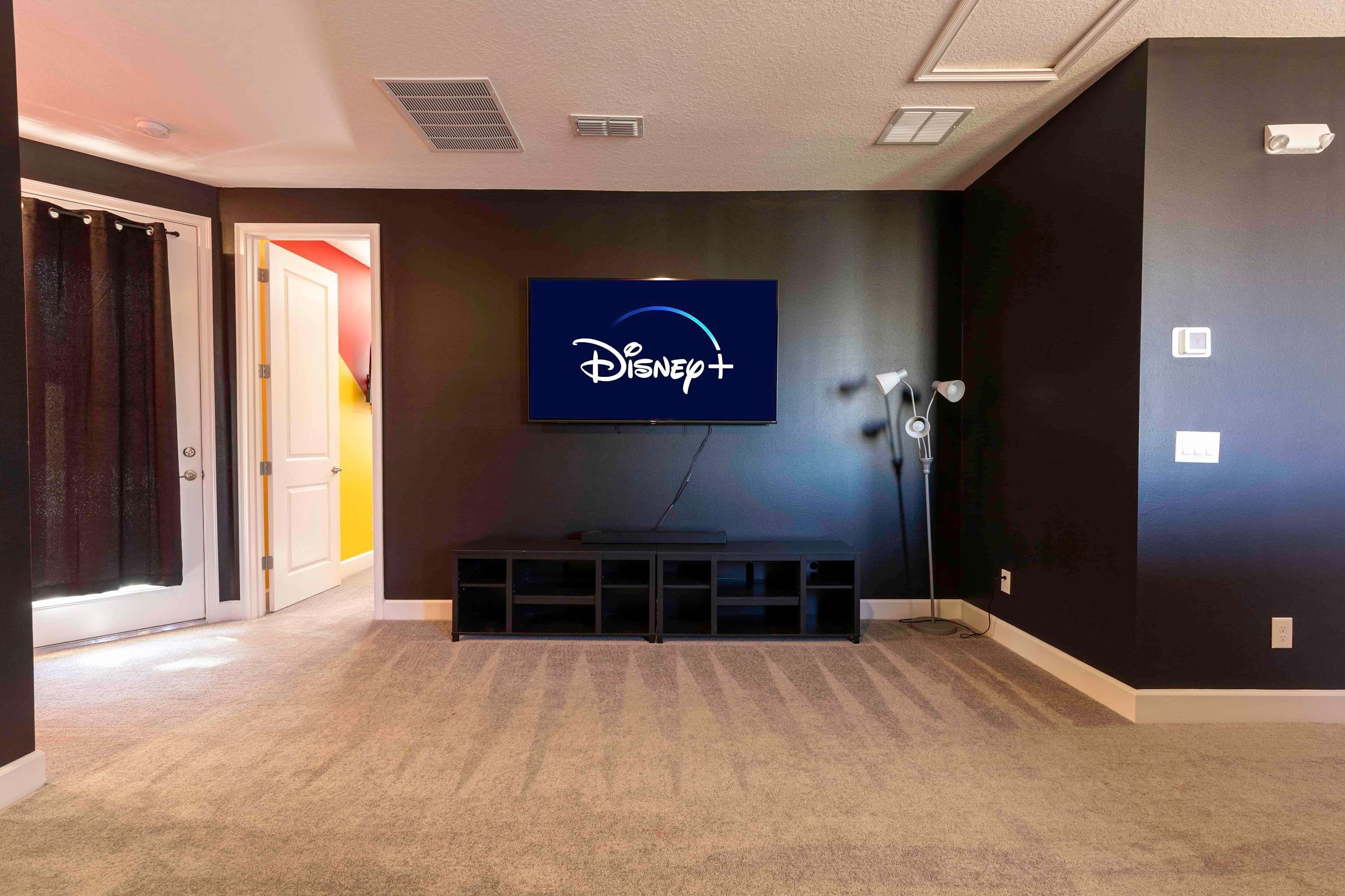 Ein Zimmer mit einem an der Wand montierten Flachbildfernseher, der das Disney+-Logo zeigt, einem dunkelgrauen Teppich und einer Tür zu einem anderen Raum.