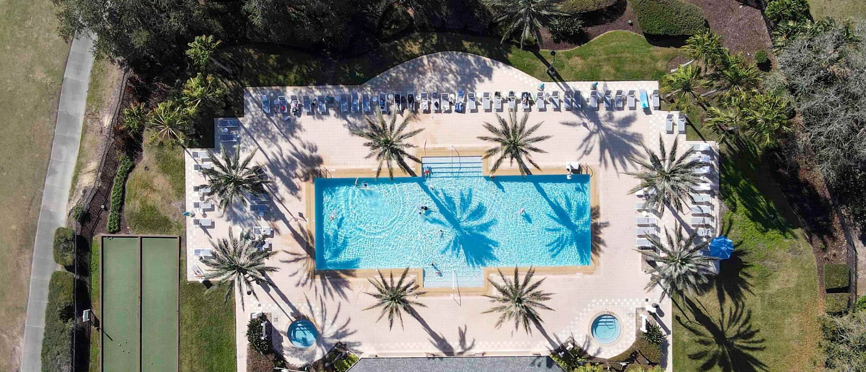 An aerial view of a resort with a large rectangular swimming pool surrounded by palm trees and lounge chairs.