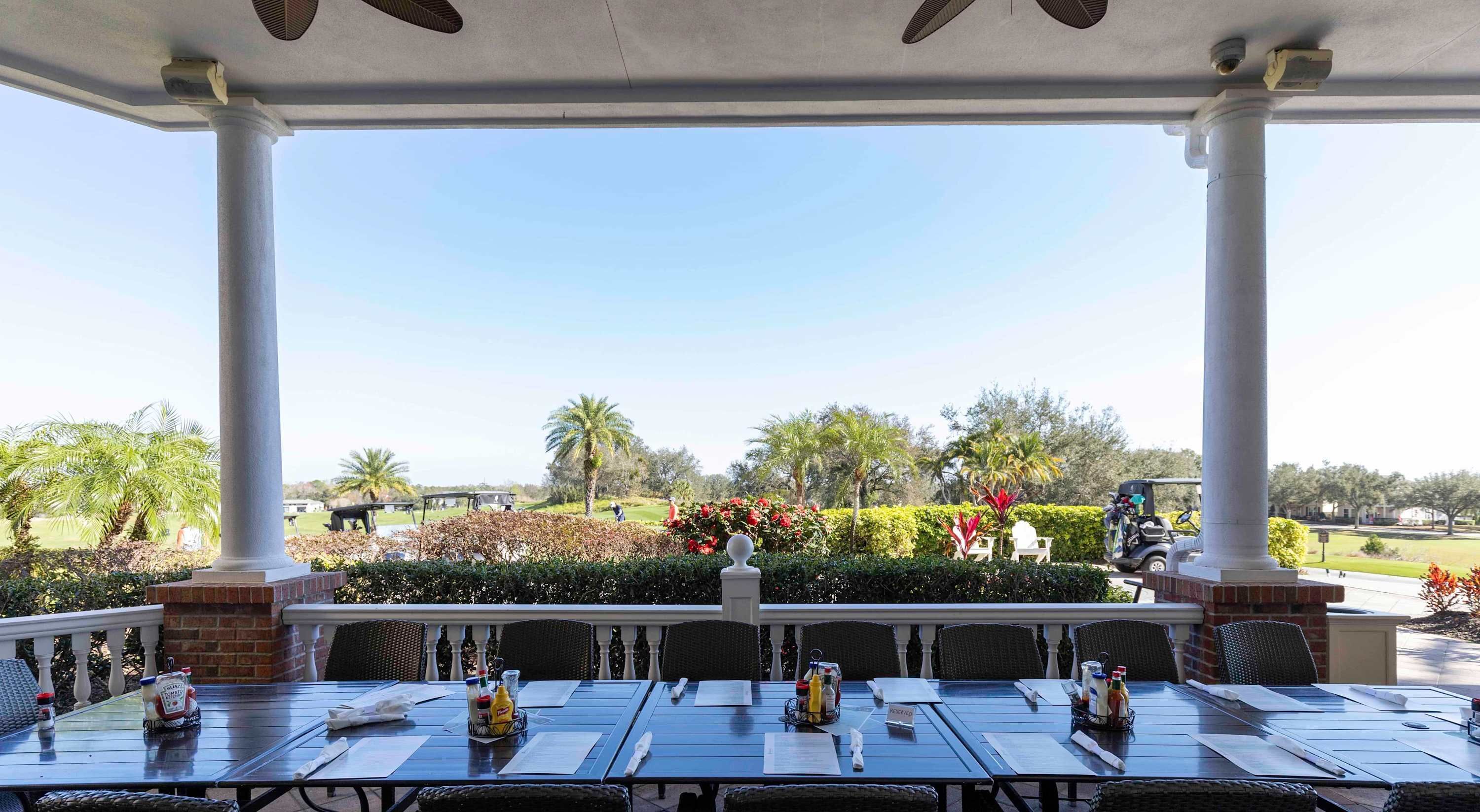Un espace de restauration extérieur abrité, avec des tables dressées, donnant sur un jardin aménagé avec des palmiers et, au loin, sur un terrain de golf.
