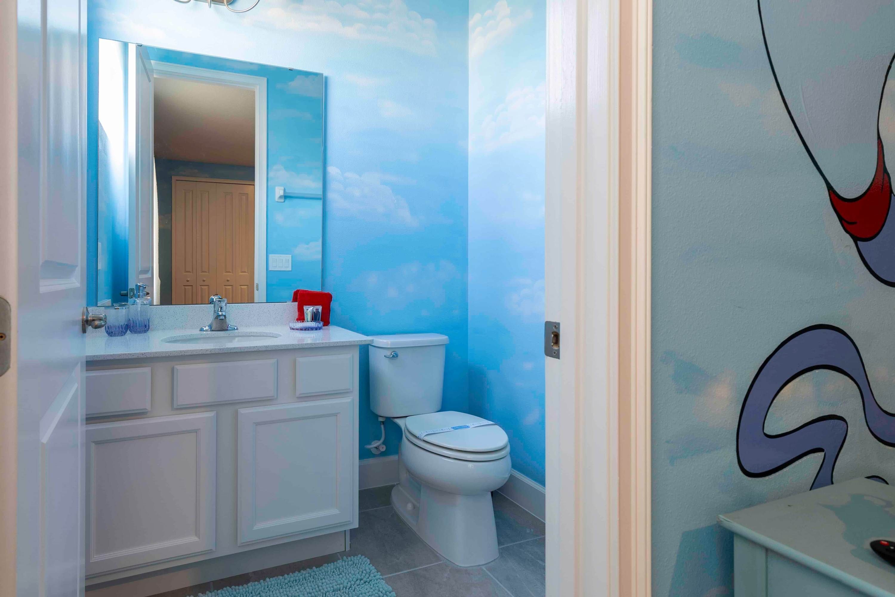 A bathroom with a blue ceiling mural, a white vanity with a mirror, a toilet, and a blue rug.
