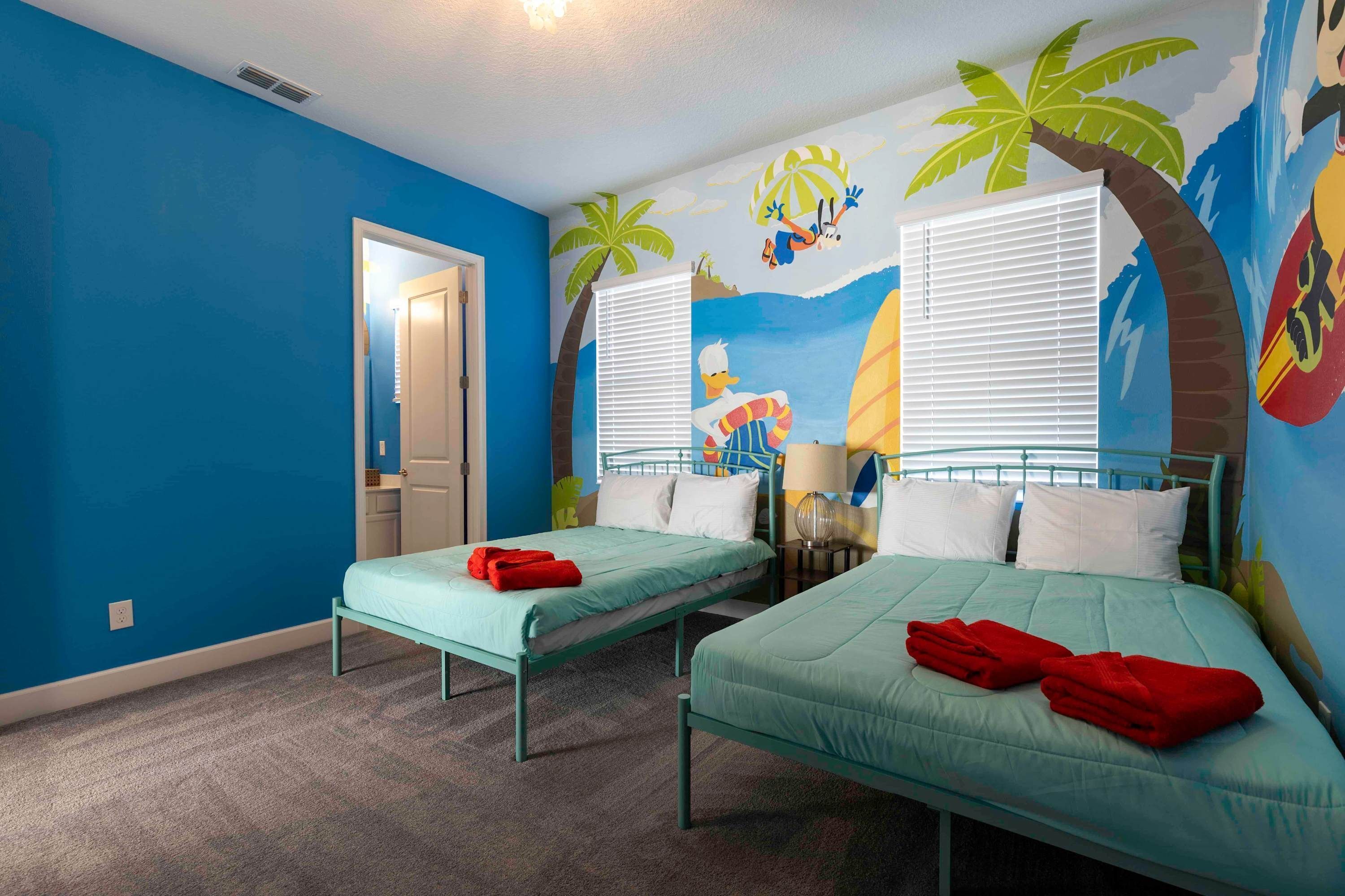 A room with two beds, each with a blue mattress and red towels. The walls are painted blue, and there is a mural of a beach scene with palm trees and a surfboard.