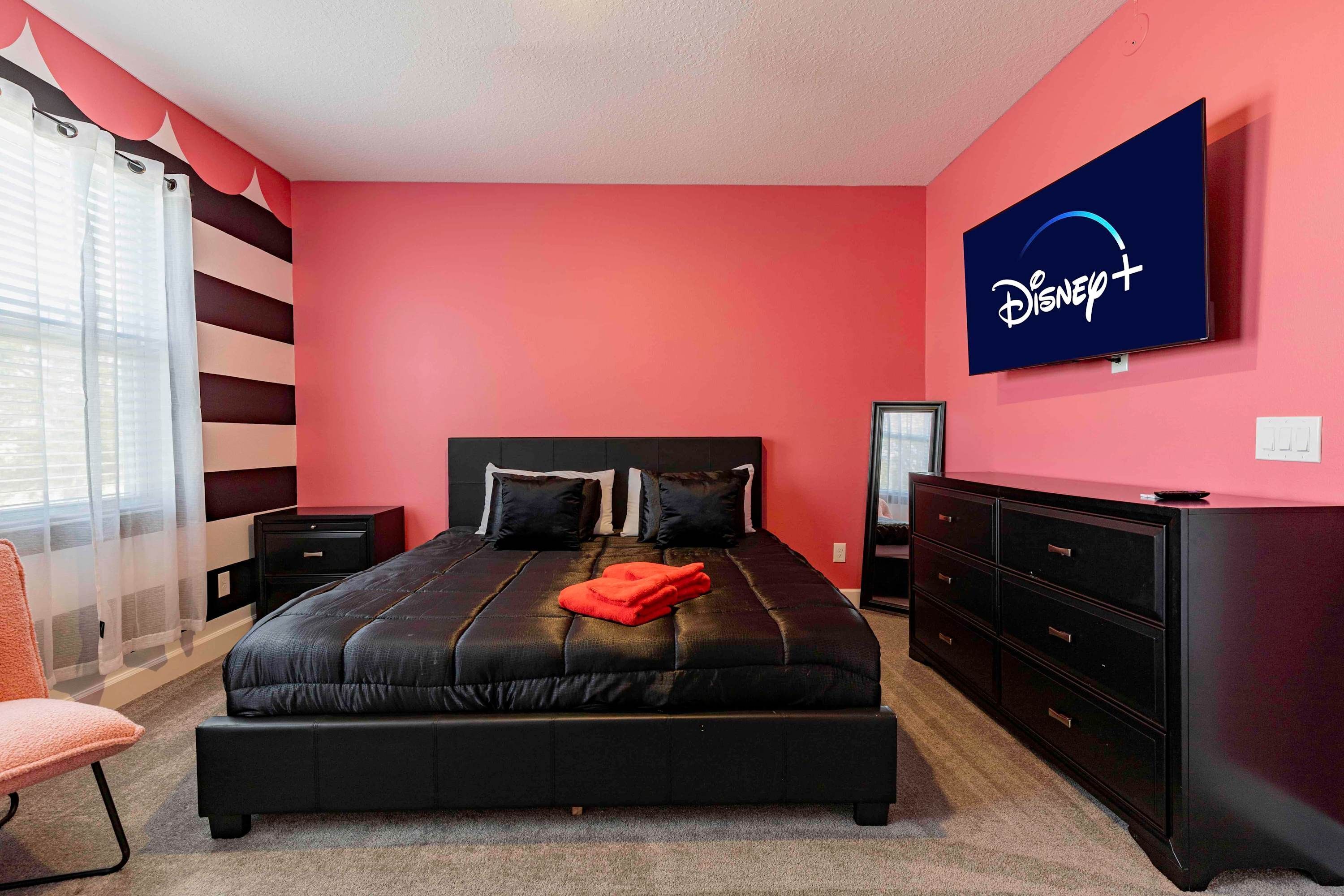 A bedroom with a large bed, a dresser, a TV mounted on the wall, and a window with curtains.