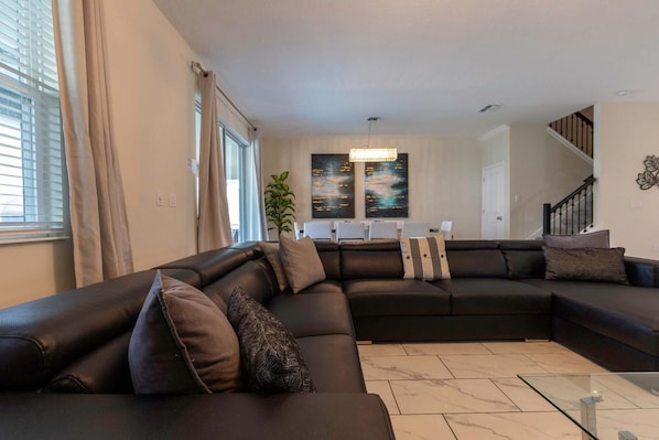 A modern living room with a large L-shaped black leather sofa, a glass coffee table, and a staircase leading to an upper floor. - Luxury 9BR Villa Poolside Bliss n Pool Table Fun (Kissimmee)
