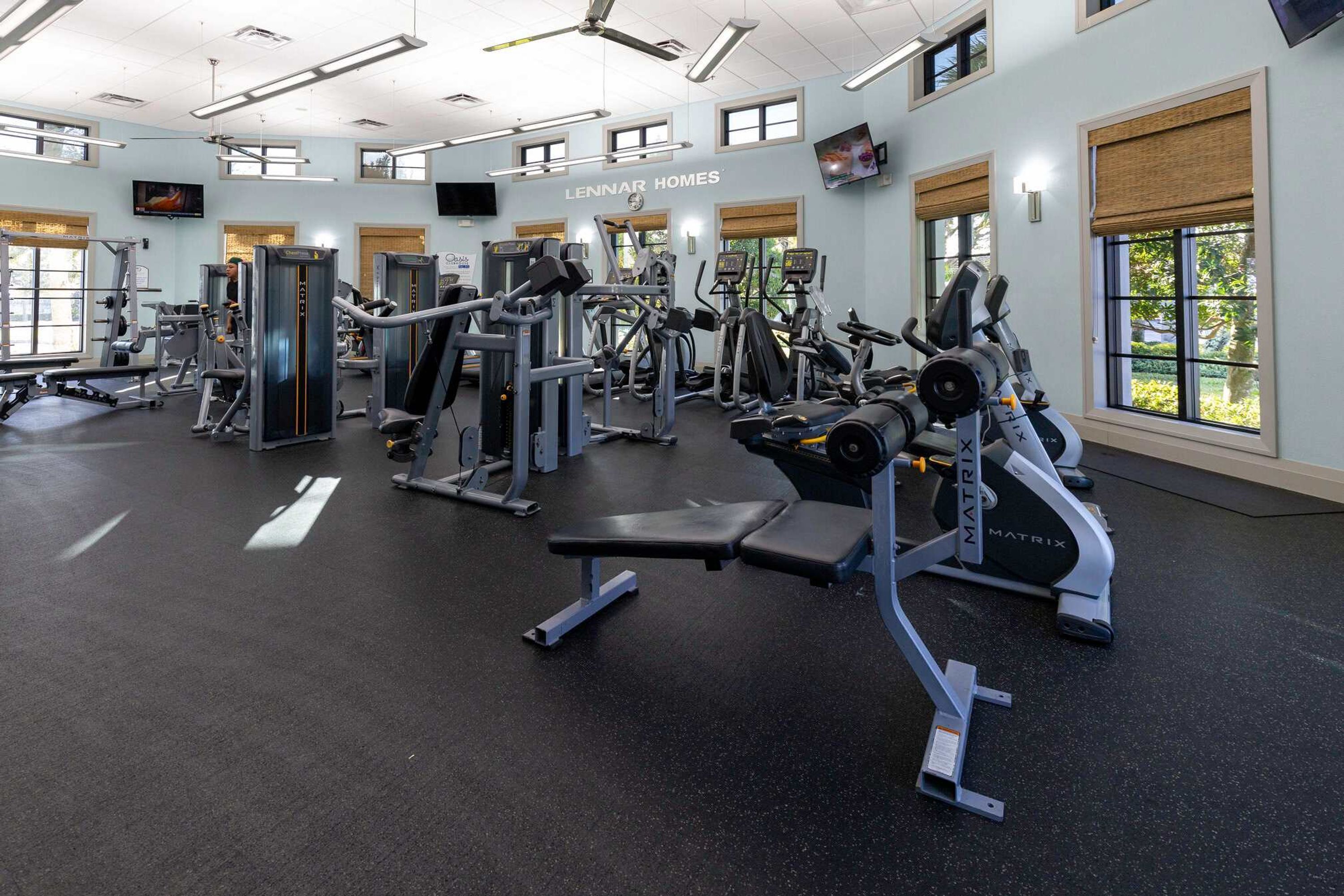 A gym with various exercise machines, a television mounted on the wall, and large windows with blinds.
