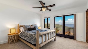 Un dormitorio con cama de madera, ventilador de techo, mesita de noche con lámpara y vistas a un balcón con paisaje panorámico.