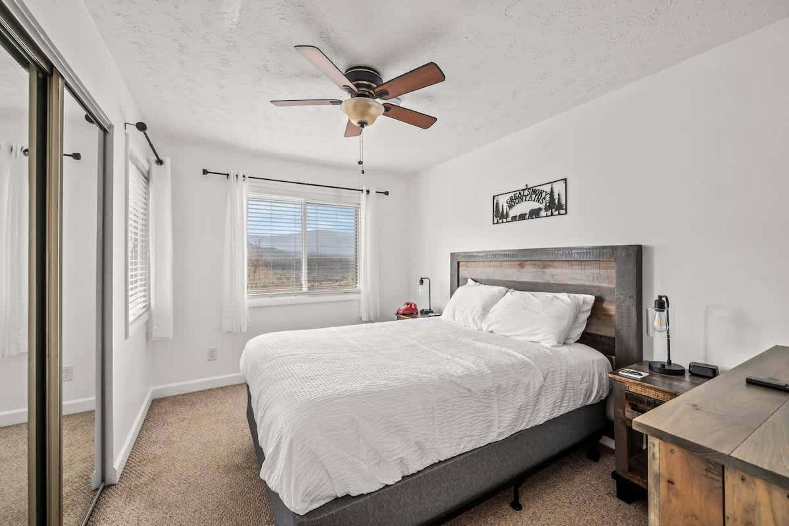 A bedroom with a bed, a ceiling fan, a window with blinds, a bedside table with a lamp, and a wall-mounted artwork.