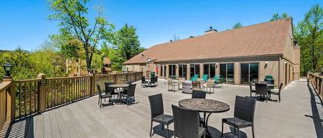 An outdoor patio area with multiple tables and chairs, a wooden deck, and a building with large windows in the background.