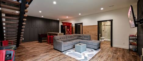 A spacious living area with a large gray sectional sofa, a wooden staircase, and a kitchen area with a red accent wall.