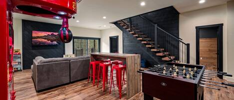 A modern living room with a sofa, red bar stools, a foosball table, and a staircase.
