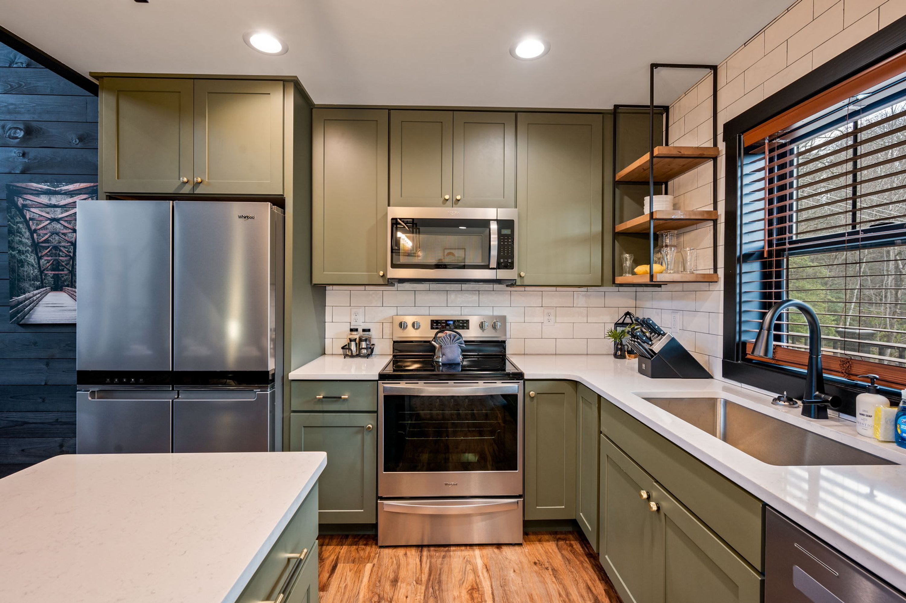 A modern kitchen with stainless steel appliances, green cabinets, and a window with blinds.