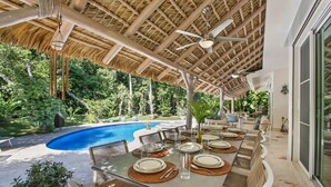 Un área de comedor al aire libre con piscina, cubierta por un techo de paja y rodeada de exuberante follaje.