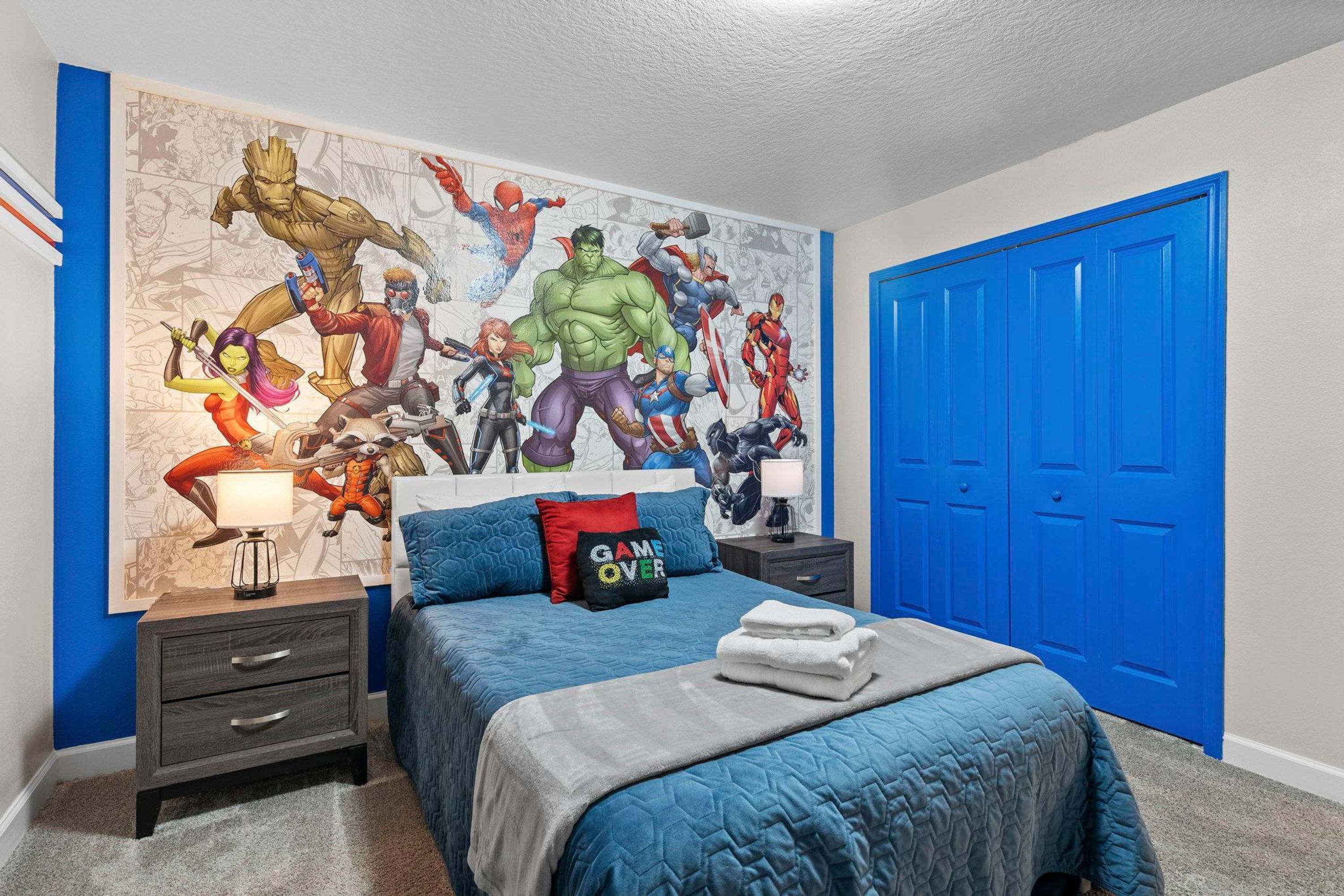 Een slaapkamer met een grote muurschildering met superhelden, een blauwe deur en een net opgemaakt bed met een 'GAME OVER' kussen.