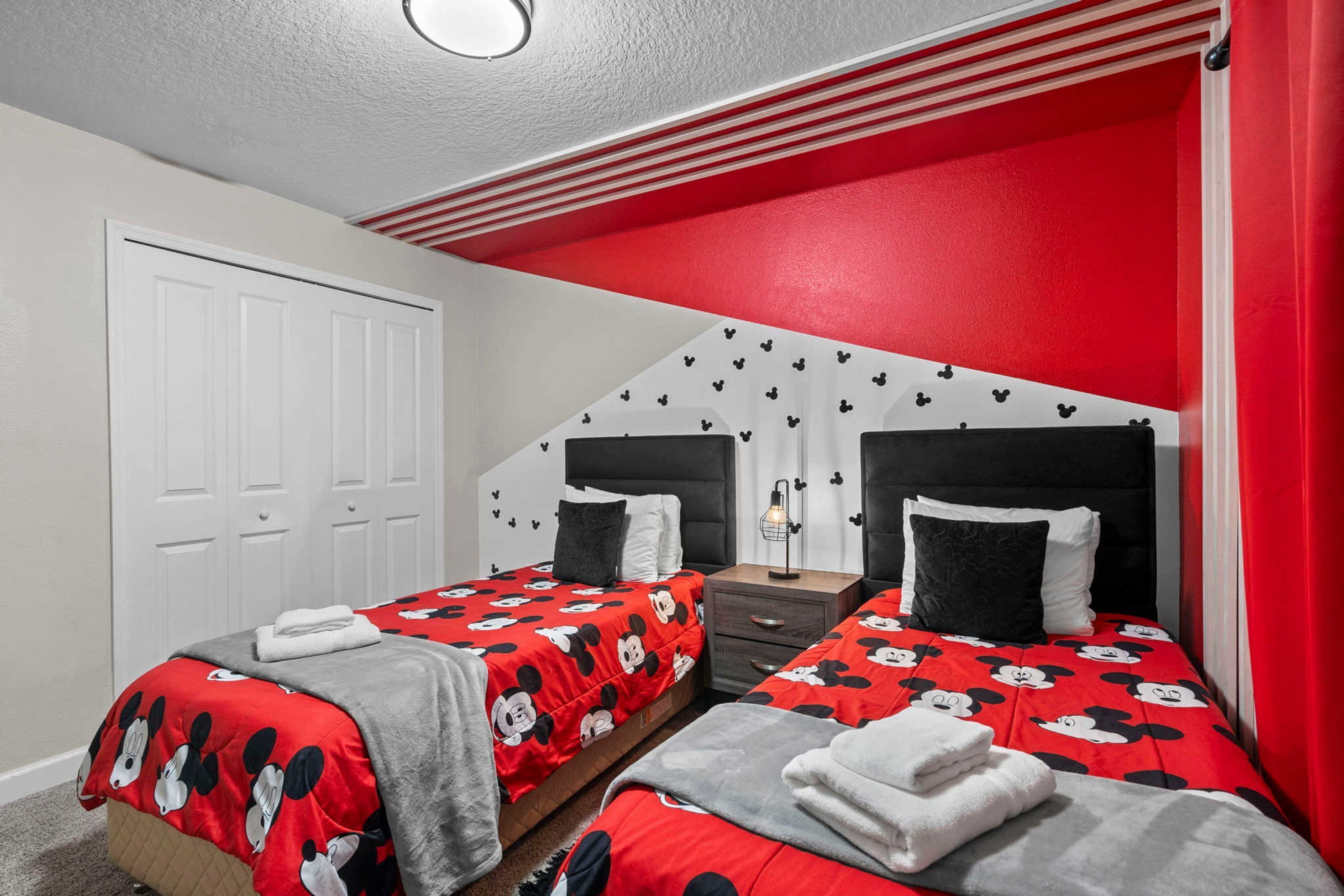 Une chambre d’hôtel avec deux lits, chacun agrémenté de linge de lit aux motifs de Mickey Mouse, une porte blanche et des touches de couleur rouge sur les murs et le plafond.