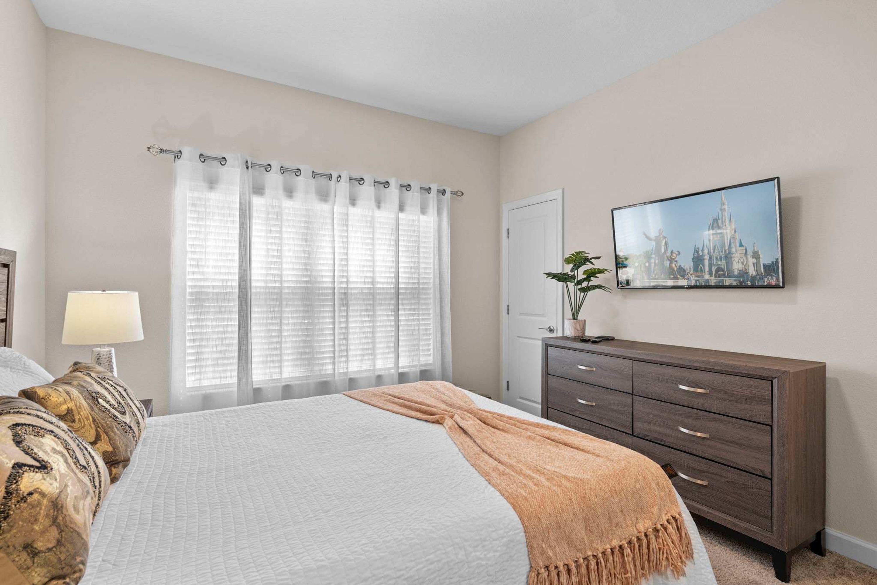 A bedroom with a bed, a dresser, a lamp, a window with curtains, and a wall-mounted picture.