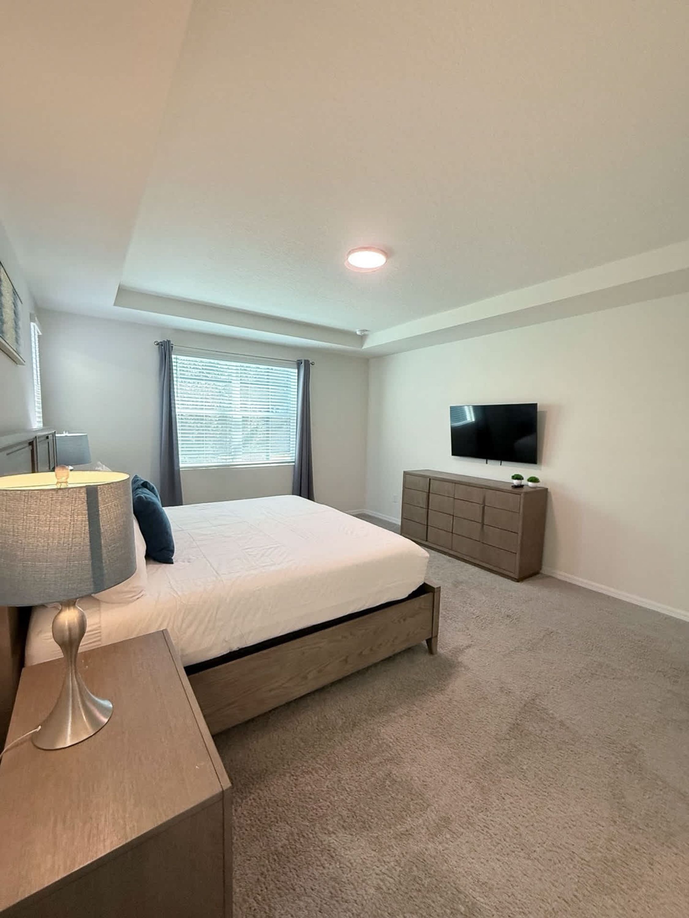 Een slaapkamer met een bed, een televisie, een dressoir, een lamp en een raam met gordijnen.