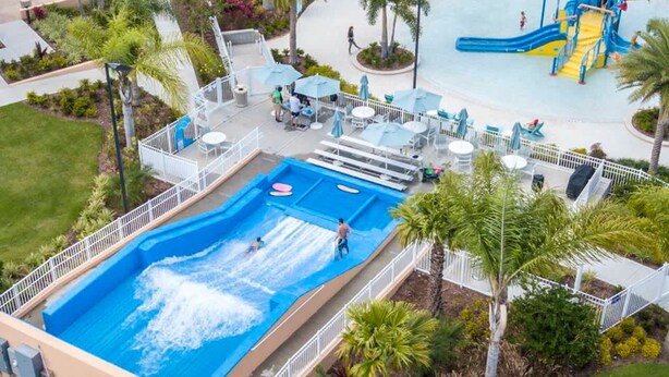 An aerial view of a water park with a large pool, slides, and lounge areas surrounded by palm trees.