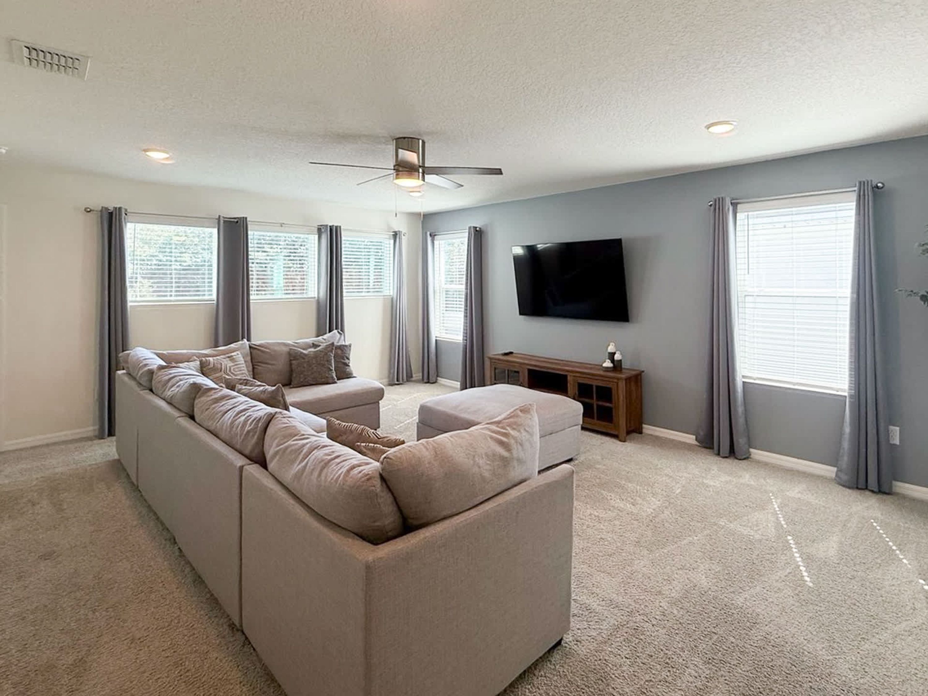 A spacious living room with a large sectional sofa, a flat-screen TV mounted on the wall, and a ceiling fan.