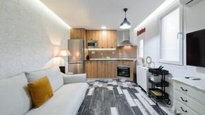 A compact kitchen with wooden cabinets, a microwave, a stove, and a sink. There is a white sofa with a mustard-colored pillow, a small table with a lamp, and a TV mounted on the wall. - GuestReady - A lovely home near Plaza Mayor (Madrid)