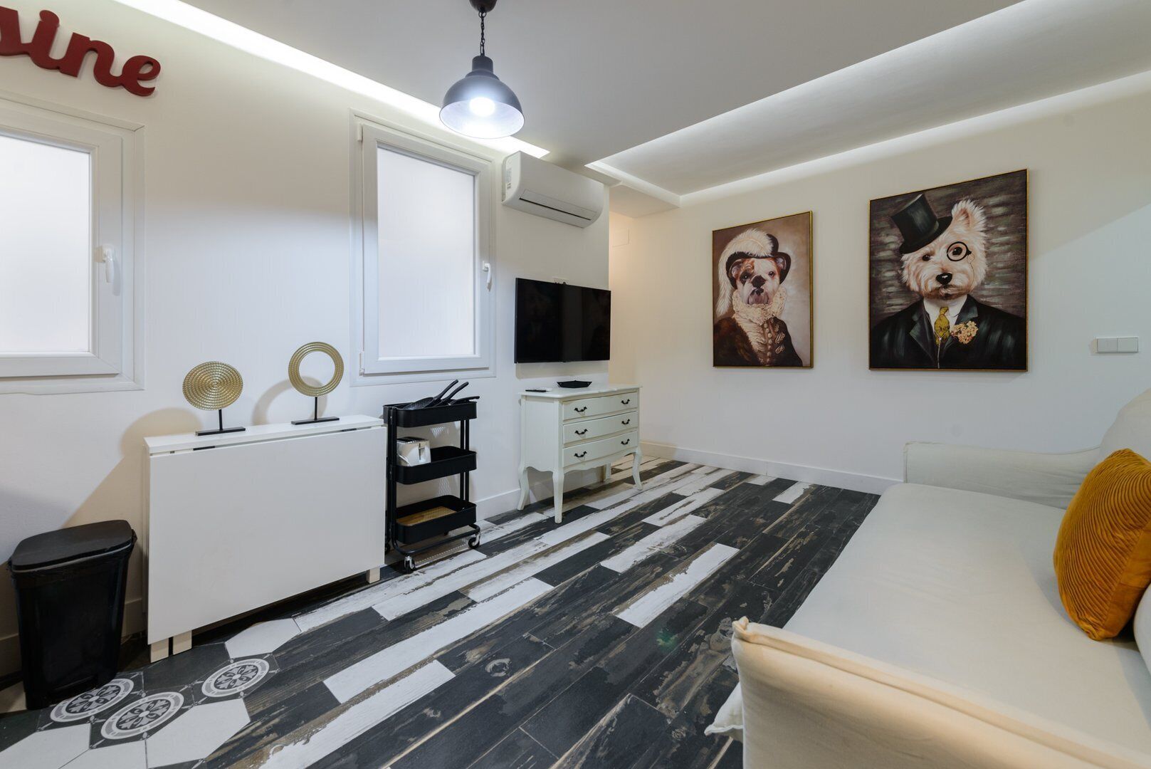 A modern bedroom with a black and white patterned floor, a white bed with a yellow pillow, a desk with a TV, and two wall-mounted paintings of dogs.