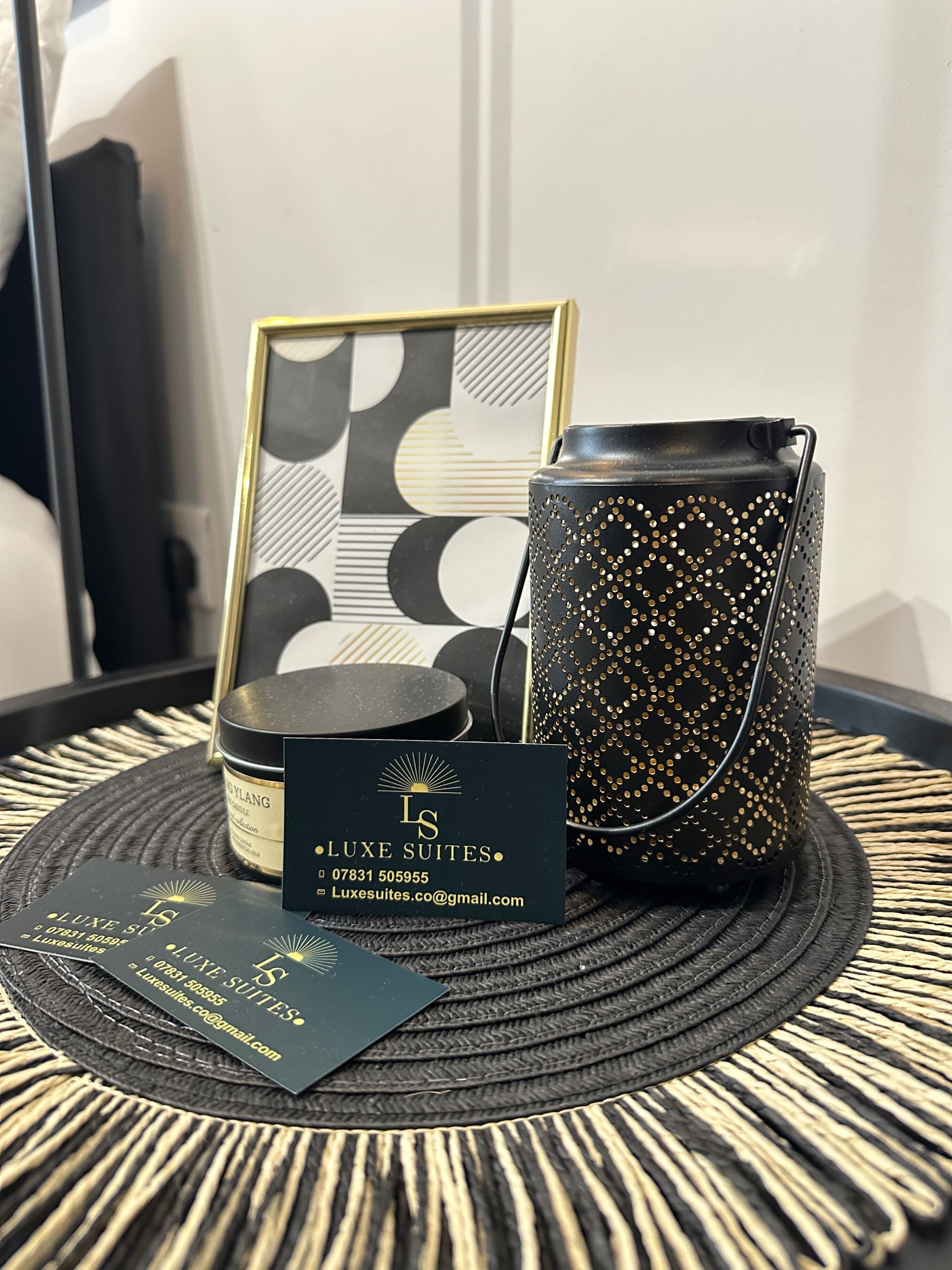 Un vase noir et doré, un tableau encadré et une carte noire portant l’inscription « LUXE SUITES » sur une table en osier.