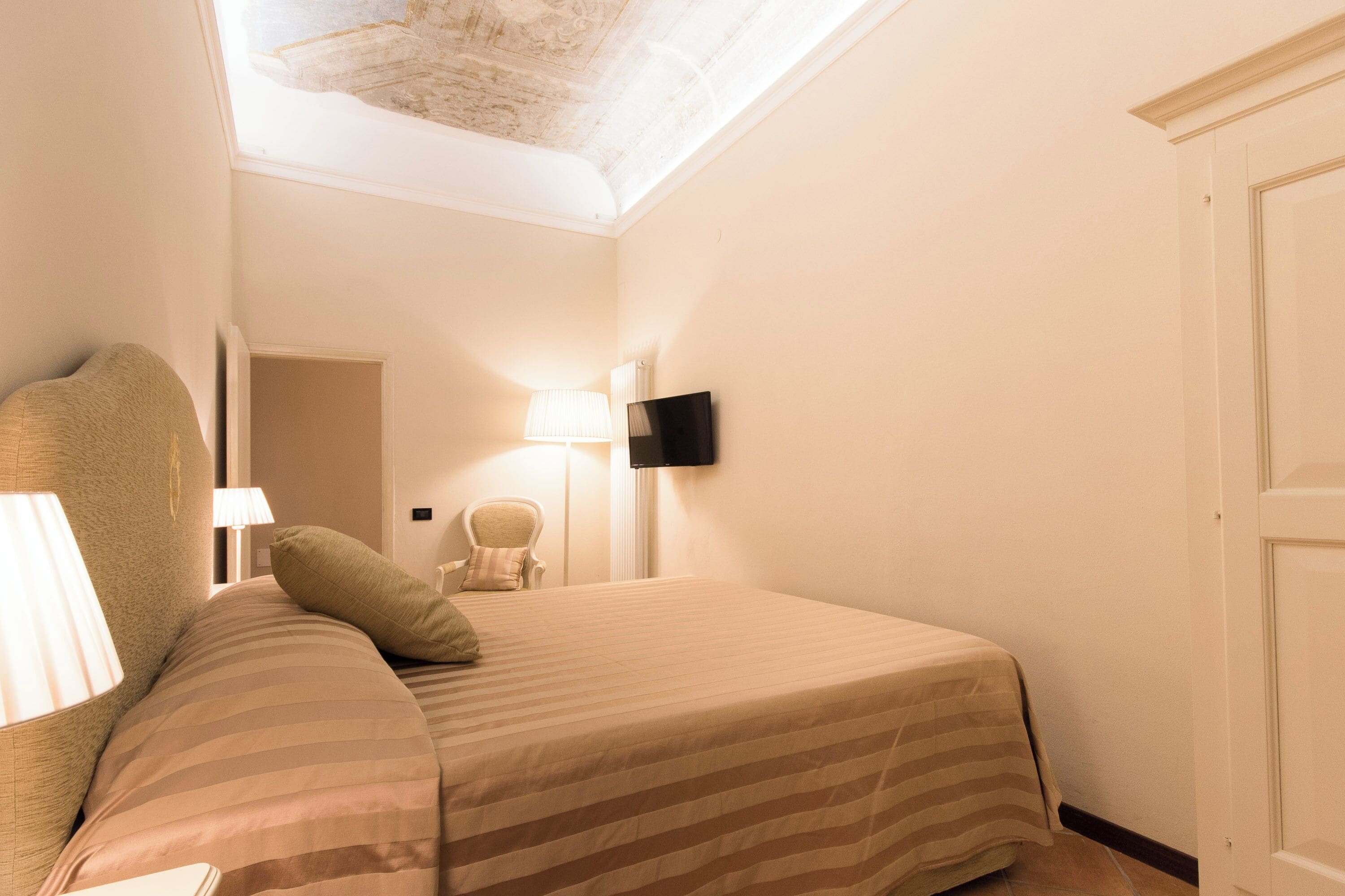 Una camera d'albergo con un letto, una sedia, una TV a parete e una lampada.