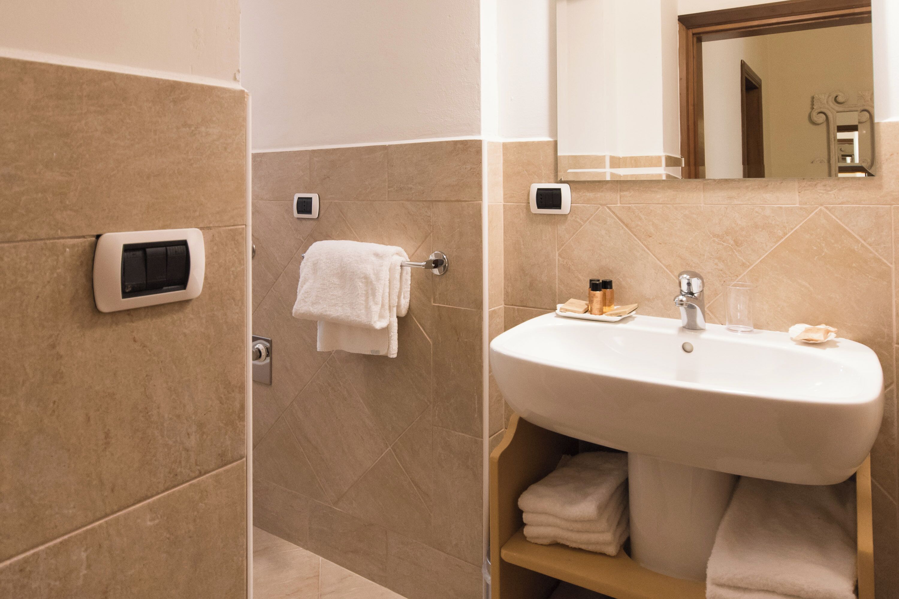 Un baño con pared de azulejos color beige, un lavamanos blanco con espejo arriba y un perchero con una toalla blanca.