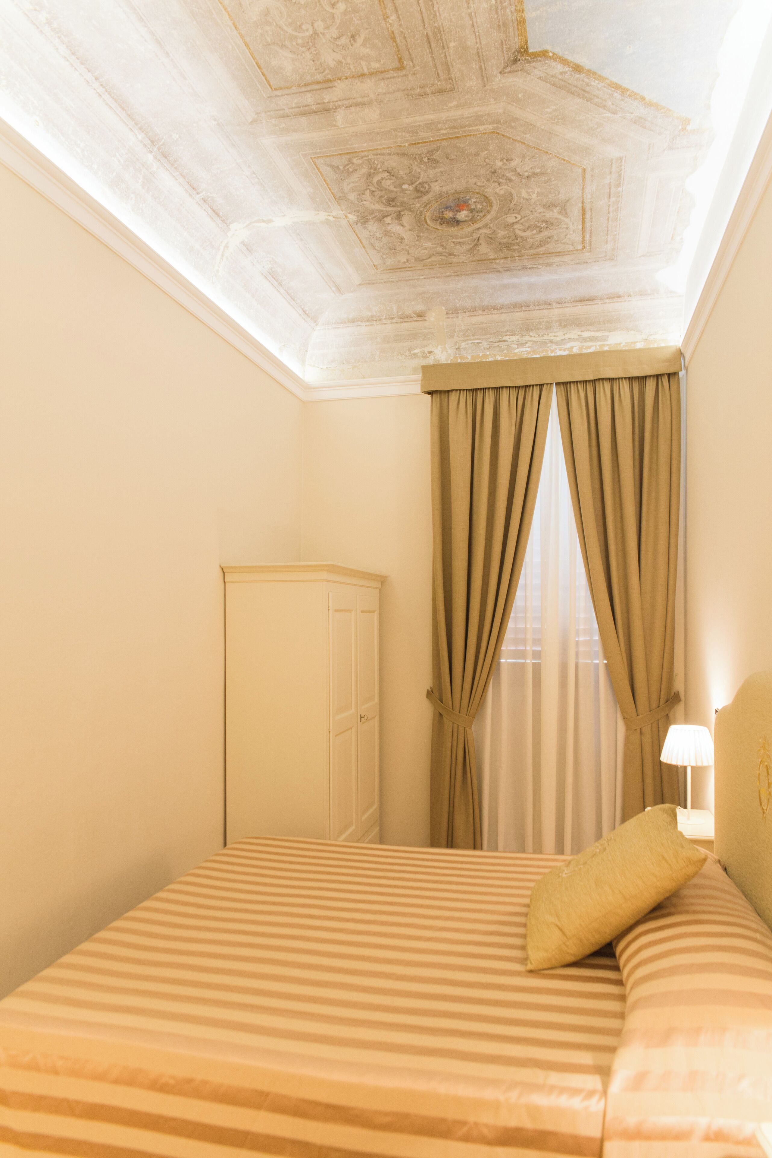 A hotel room with a bed, a wardrobe, a window with curtains, and a ceiling with a decorative design.