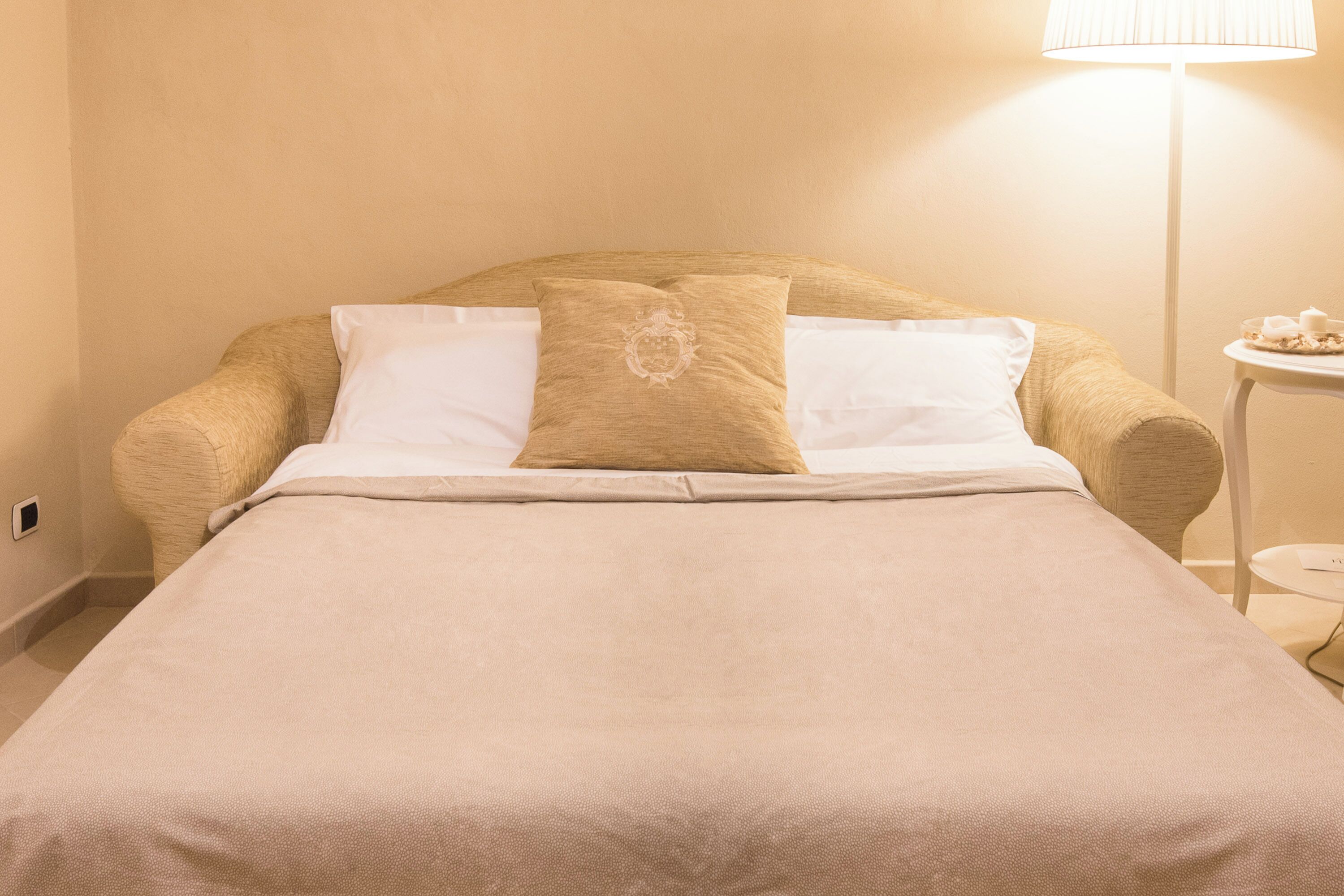A neatly made bed with a beige bedspread and two pillows, one with a decorative emblem, in a room with a lamp and a small table.