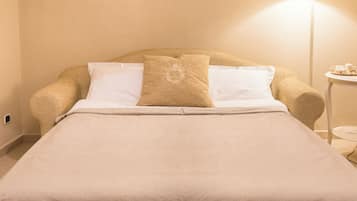 A neatly made bed with a beige bedspread and two pillows, one with a decorative emblem, in a room with a lamp and a small table.