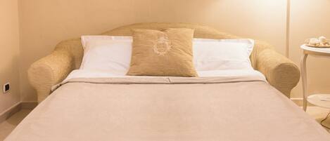 A neatly made bed with a beige bedspread and two pillows, one with a decorative emblem, in a room with a lamp and a small table.