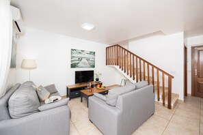 A modern living room with a grey sofa set, a wooden coffee table, a flat-screen TV, and a staircase with wooden railings.
