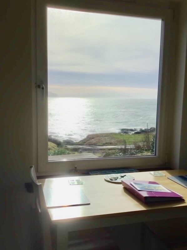 A room with a large window overlooking the sea, a desk with a notebook and a book, and a view of rocky coastline. - Luxury Apartment in absolutely stunning seafront location, Isle of Skye. (Isle of Skye)
