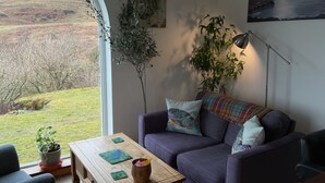 A modern living room with a purple sofa, a wooden coffee table, and a view of a grassy landscape through a large window.